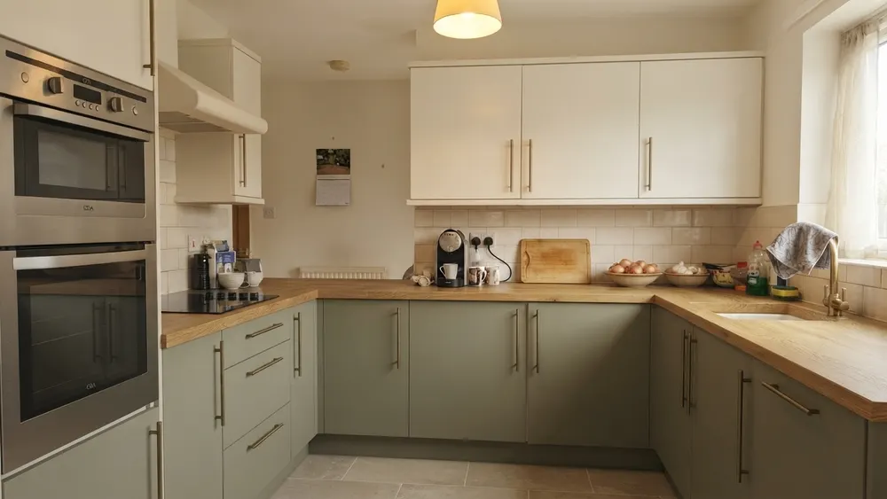 A kitchen with cream upper cabinets, sage green lower cabinets, a warm oak countertop, and the original tile backsplash kept in place
