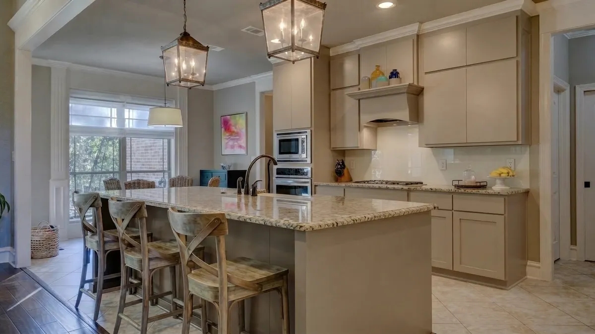 Open kitchen with greige cabinets, speckled light granite countertops and island, and a cream back-painted glass backsplash under a wood range hood, with three wooden bar stools at the island