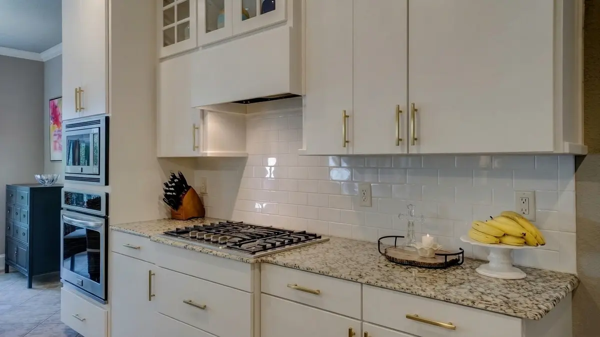 White flat-panel kitchen cabinets with brass bar pulls, a speckled light granite countertop, white subway tile, a gas cooktop and a white wall oven column, with a small dark dresser at the far left and bananas on a cake stand