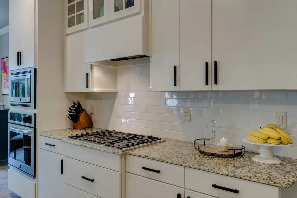 The same white kitchen with slim matte black bar pulls on every door and drawer, speckled granite countertop, white subway tile, gas cooktop and a black wire tray with a candle on the counter