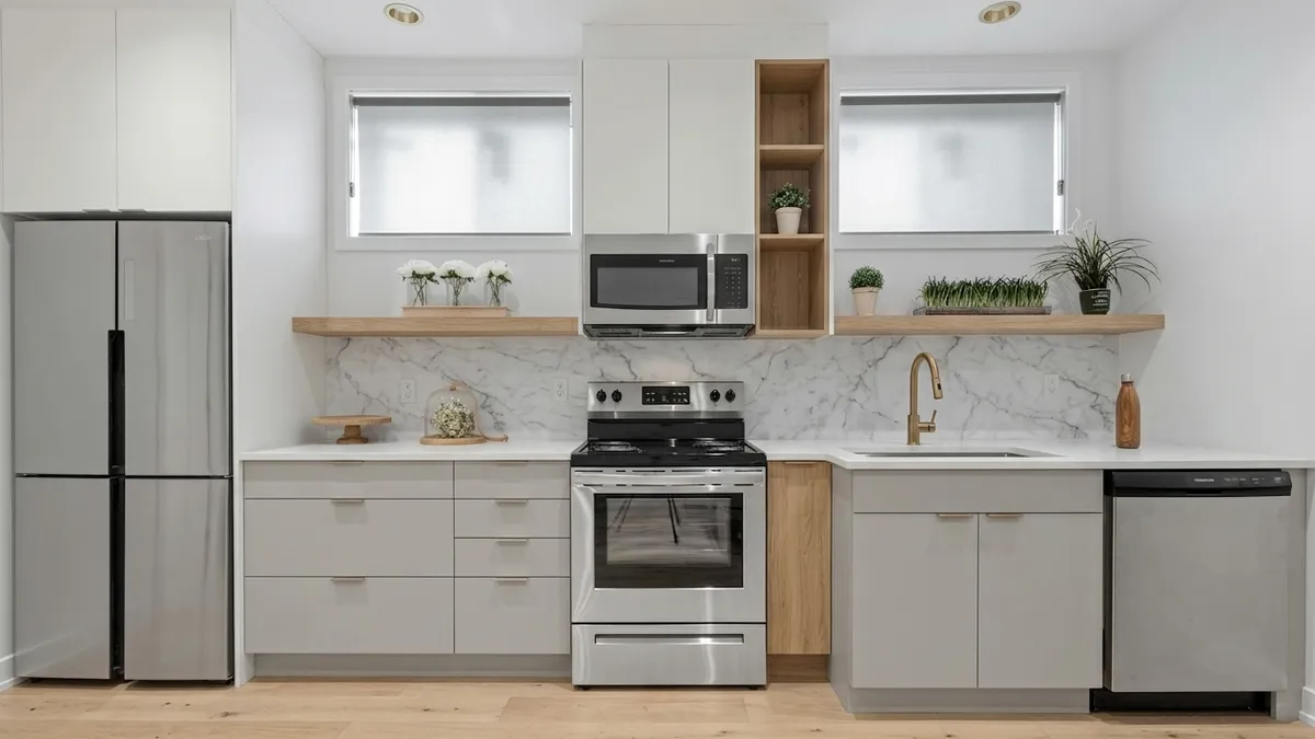 A straight-run kitchen redesigned with flat light gray cabinet fronts, a white countertop, a marble-look backsplash, a brass-toned faucet and an open oak shelf unit beside the microwave
