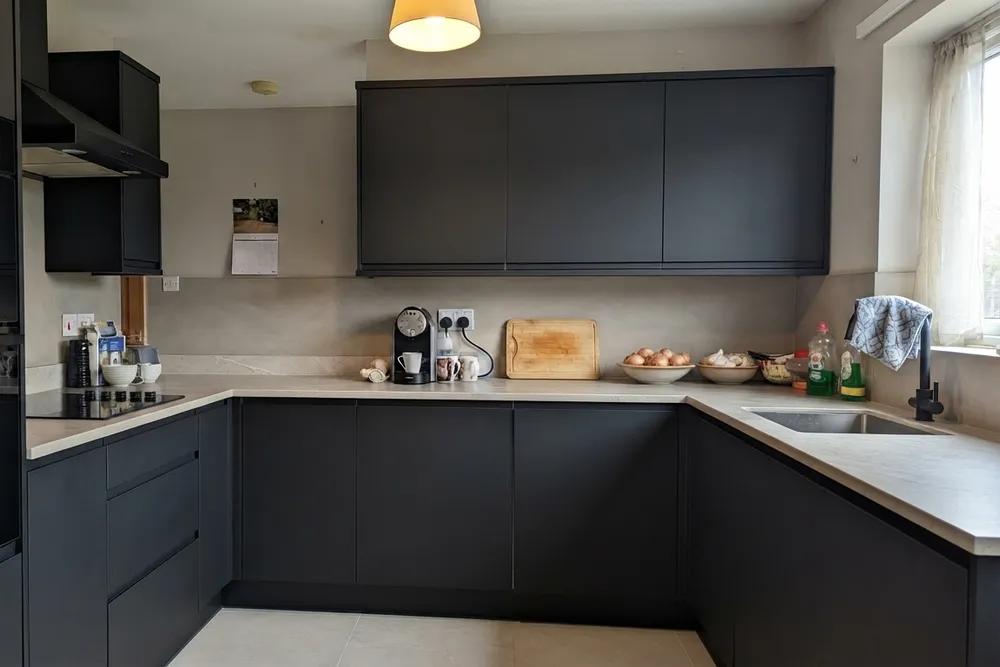 A U-shaped kitchen with matte black handleless cabinet fronts, black built-in ovens and hood, a pale stone-look countertop and backsplash, a black tap and light floor tiles