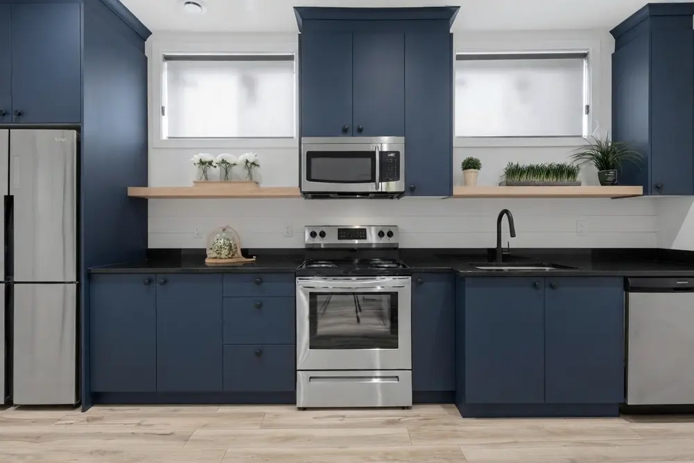 The same kitchen wall with flat navy cabinets, small matte black round knobs, a polished black granite countertop, the white shiplap backsplash and light wood open shelves