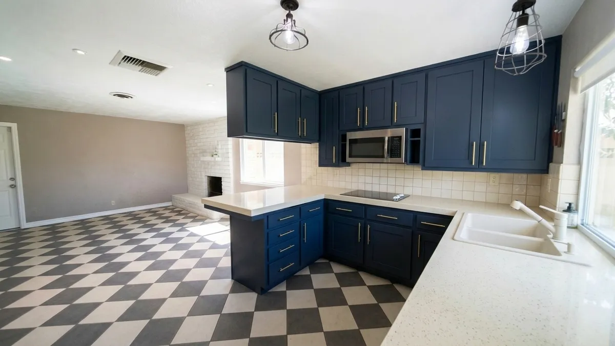A kitchen with navy shaker cabinets and brass bar pulls, a smooth cream countertop and cream square backsplash tiles, a white double-bowl sink, a black-and-white checkerboard floor and taupe walls