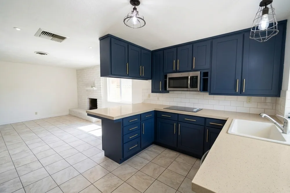 The same L-shaped kitchen with navy shaker cabinets and brass pulls, a beige speckled countertop, white subway-style tiles, a single white sink, beige square floor tiles and white walls