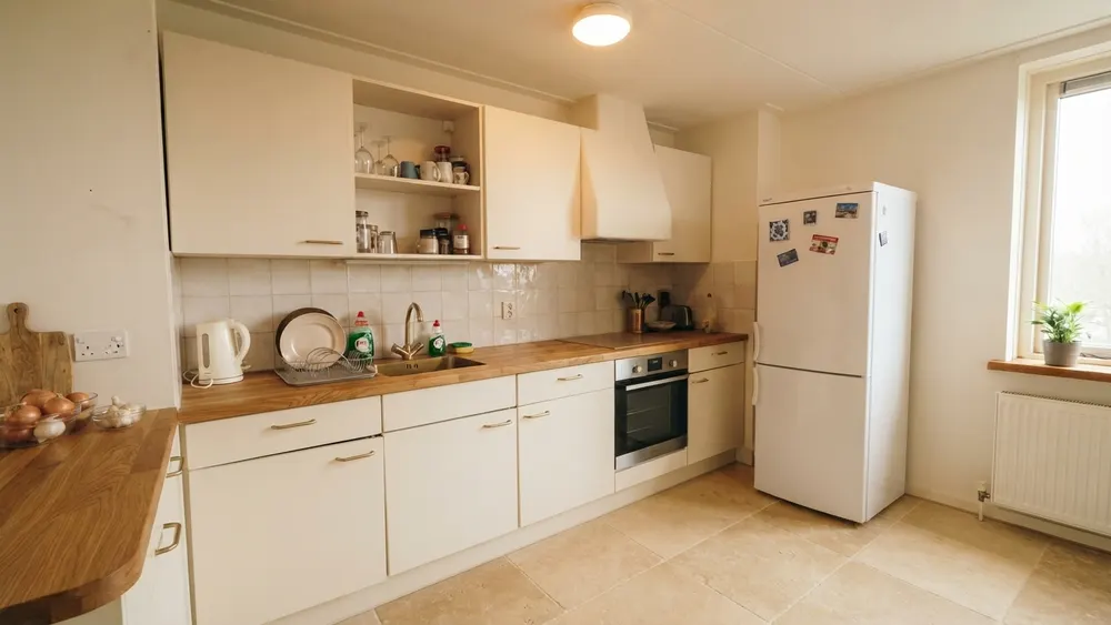 The same kitchen with cream cabinet fronts, an oak butcher-block countertop, cream tiles in the original grid, a travertine-look floor and a large cream chimney hood
