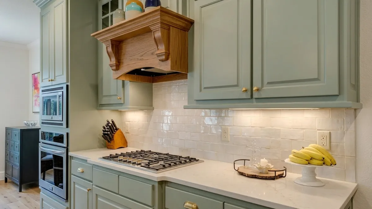 Sage green raised-panel kitchen cabinets with brass knobs, an oak range hood with corbels, glossy cream tile and a white stone-look countertop under warm under-cabinet light