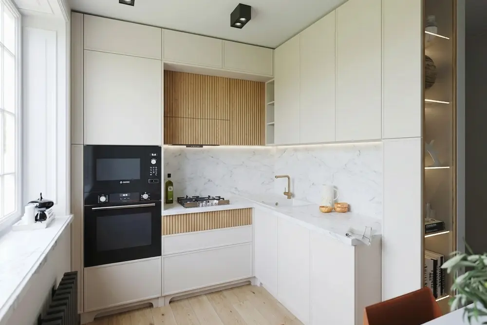 The same small kitchen with cream cabinets with a slim frame, a white marble countertop and backsplash, oak slats on the hood surround and under the cooktop, an LED strip under the uppers and a light plank floor