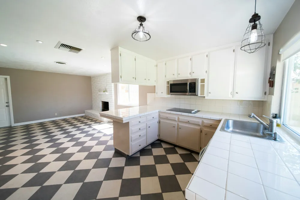 The original 1990s kitchen before any changes: cream and blush cabinets, a black-and-cream checkerboard tile floor, a white brick fireplace visible in the connected living area