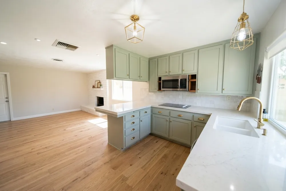 The same kitchen with sage green shaker cabinets, brass hardware and brass pendant lights, a white quartz countertop, a white tile backsplash and a warm oak plank floor