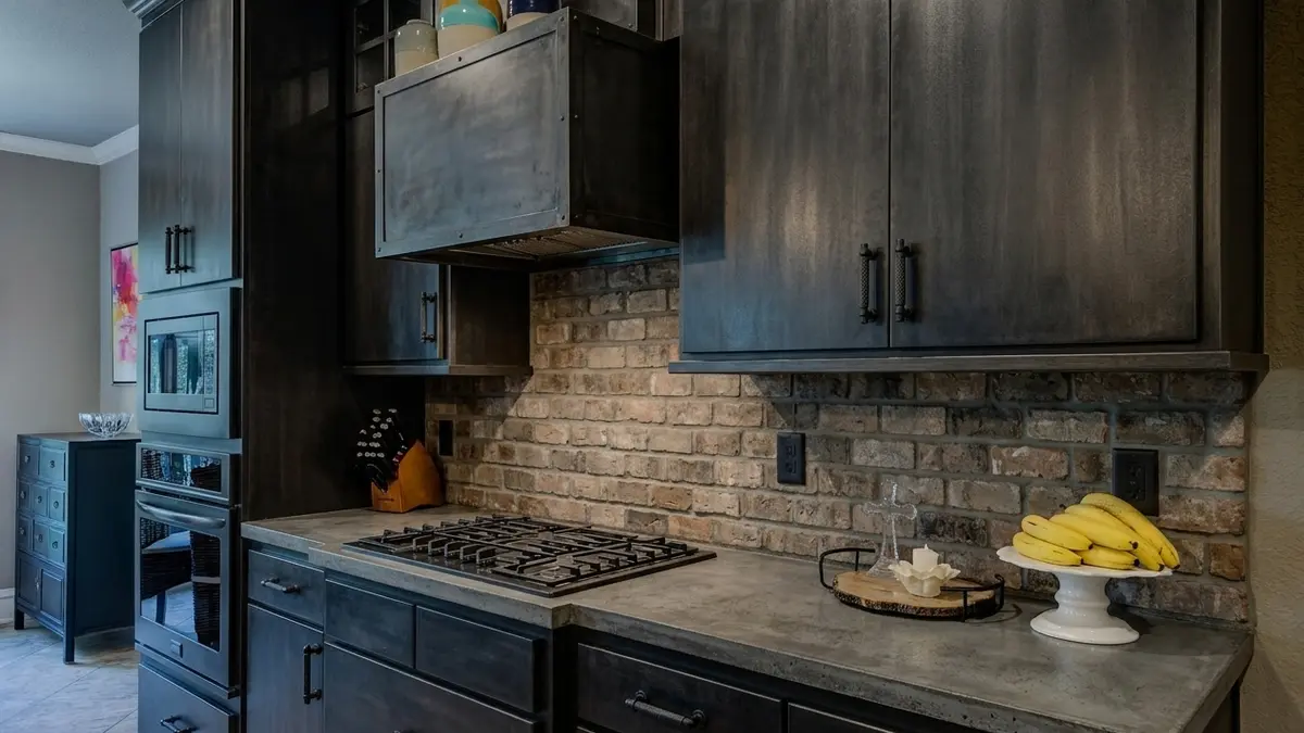 The same kitchen with dark charcoal cabinets and black bar pulls, a brick backsplash, a concrete-look countertop and a steel-clad range hood