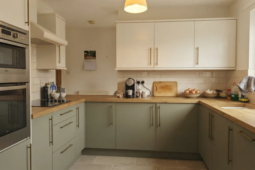 Dezelfde keuken met matte saliegroene onderkasten, mat crème bovenkasten, een eiken werkblad, messing grepen en grote grijsbeige vloertegels