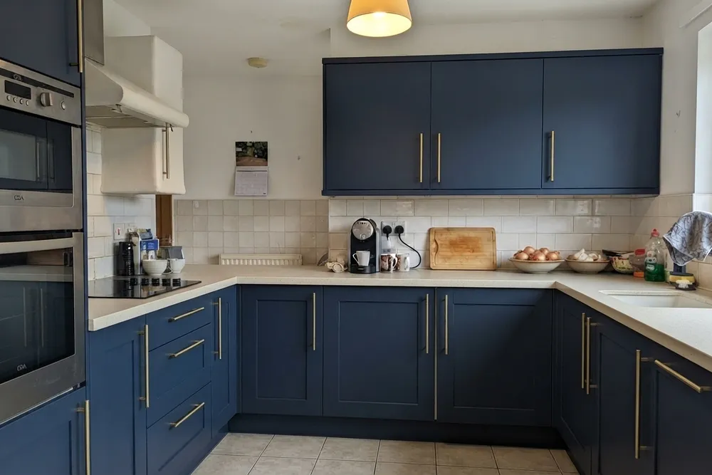 Dezelfde keuken met matte donkerblauwe kaderfronten, messing grepen, een licht gespikkeld blad en originele metrotegels