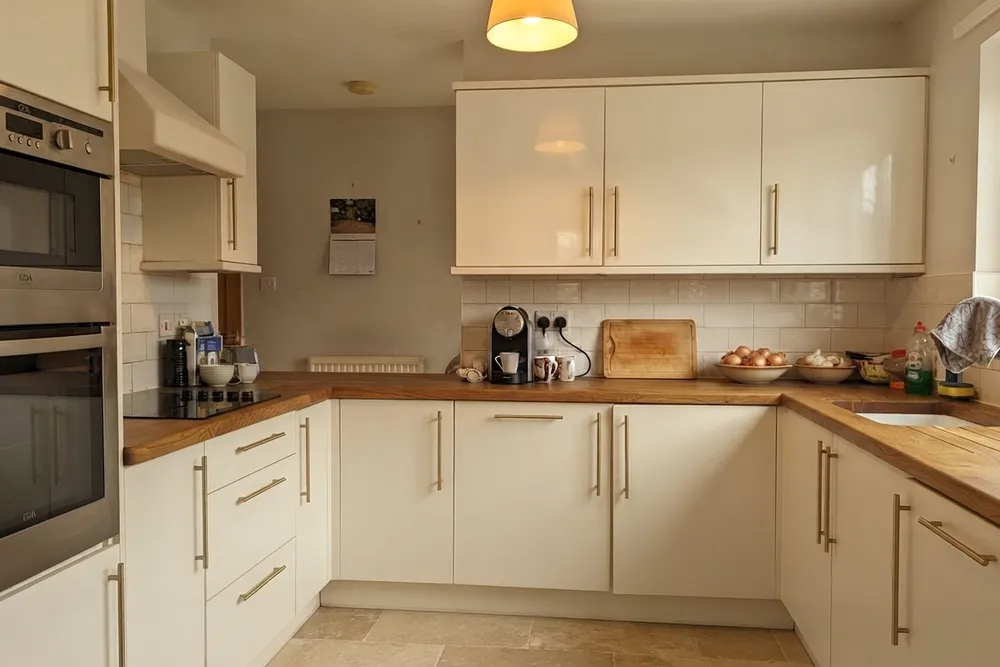 Dezelfde keuken met crème hoogglansfronten, messing balkgrepen, een eiken werkblad, een onderbouwspoelbak, beige wand en travertijnachtige vloertegels