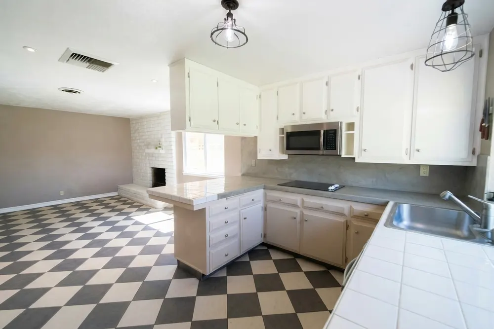 Gedateerde crème keuken met zwart-wit geblokte vloer, een achterwand en werkblad langs de wand in grijs naadloos microcement en een wit tegelblad bij de spoelbak en het schiereiland