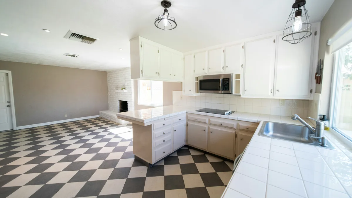 Open keuken-woonkamer met gedateerde beige kastjes, een dambordvloer in zwart-wit en een witgesausde open haard op de achtergrond